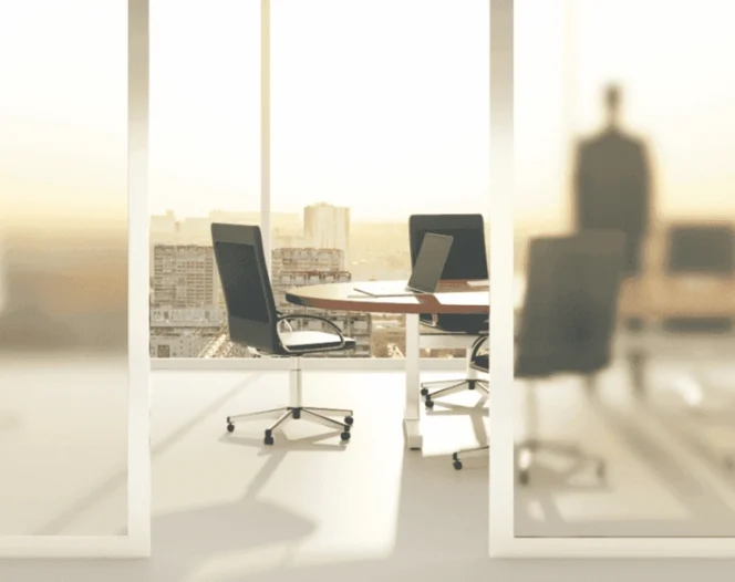 An empty meeting room with a large window overlooking a cityscape at sunset, with a person standing in the background.